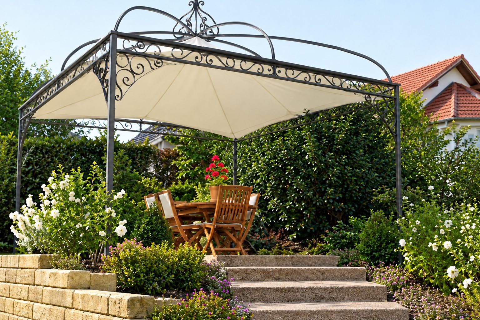 Pergola en fer forgé à toile claire dans un jardin fleuri, avec table et chaises en bois, muret de pierre et escalier au premier plan.