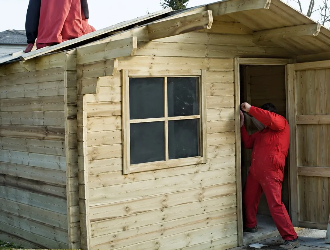 Une personne vêtue de rouge se tient à la porte d'un petit abri de jardin en bois avec une fenêtre, pendant sa construction.