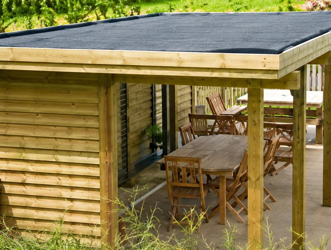 Un abri de jardin en bois à toit plat abrite une table et des chaises, posé sur une terrasse près d'une pelouse.