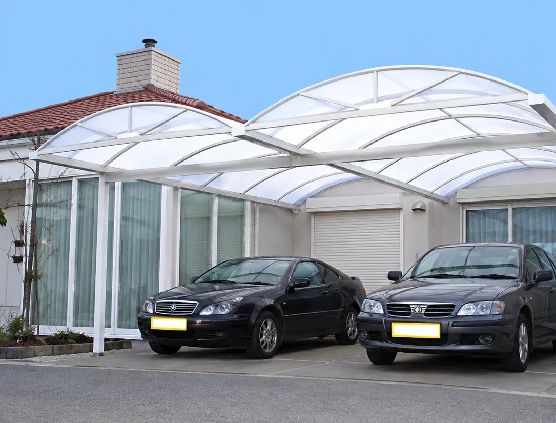Deux voitures sombres sont garées sous un grand abri translucide aux formes courbes devant une maison blanche.