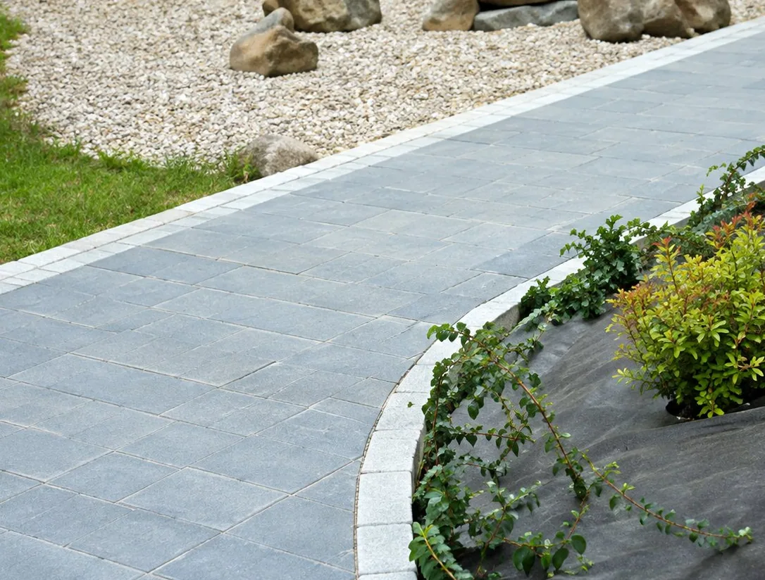 Une allée en béton gris bordée de pierres et d'un massif de plantes, avec du gravier et de la pelouse en arrière-plan.
