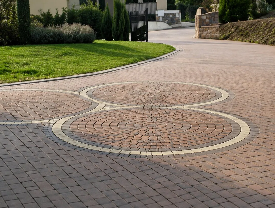 Une allée pavée de briques rouges et grises avec des motifs en forme de cercles longe une pelouse verte dans un quartier résidentiel.