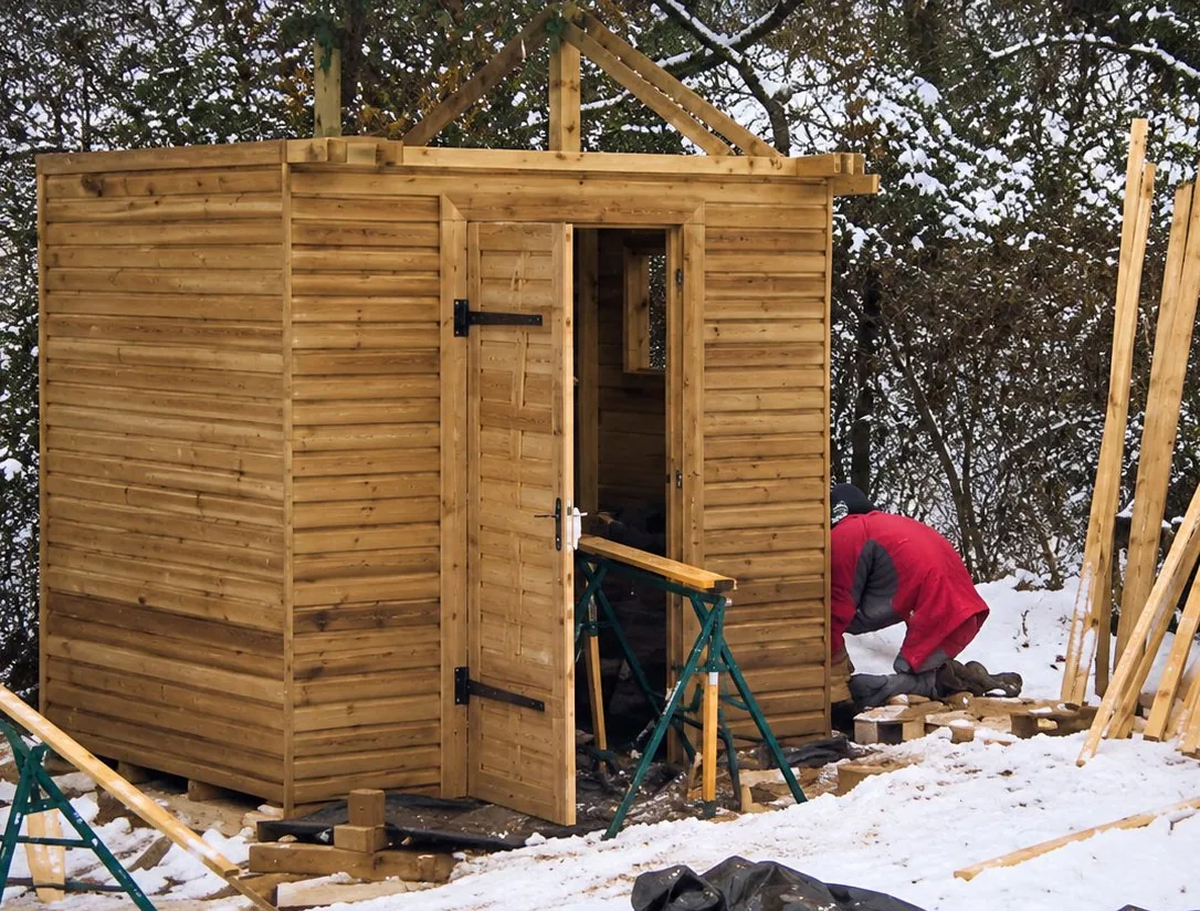 Une petite cabane en planches de bois en cours de construction sur un terrain couvert de neige, avec une personne penchée à côté et des poutres appuyées sur le côté.