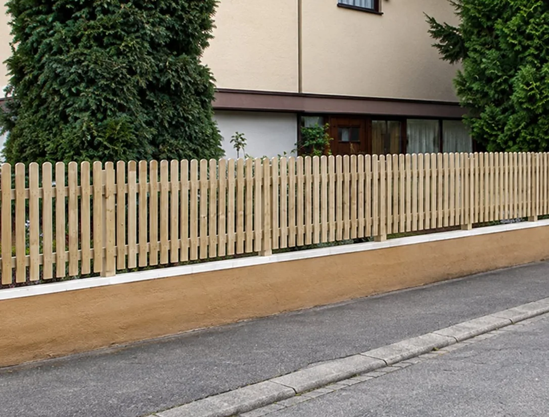 Une clôture en bois à lattes verticales longe un trottoir devant une maison à façade beige et des arbustes verts.
