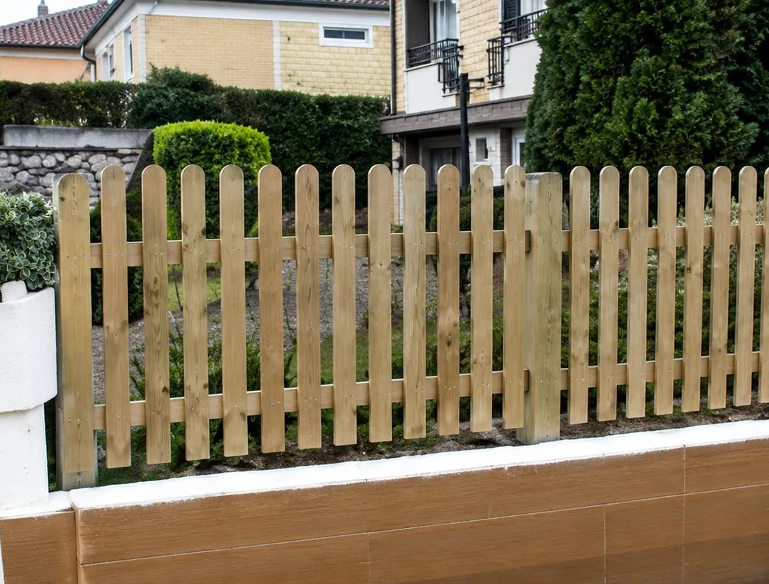 Une clôture en bois à lattes verticales posée sur un muret, devant une haie et des maisons en arrière-plan.