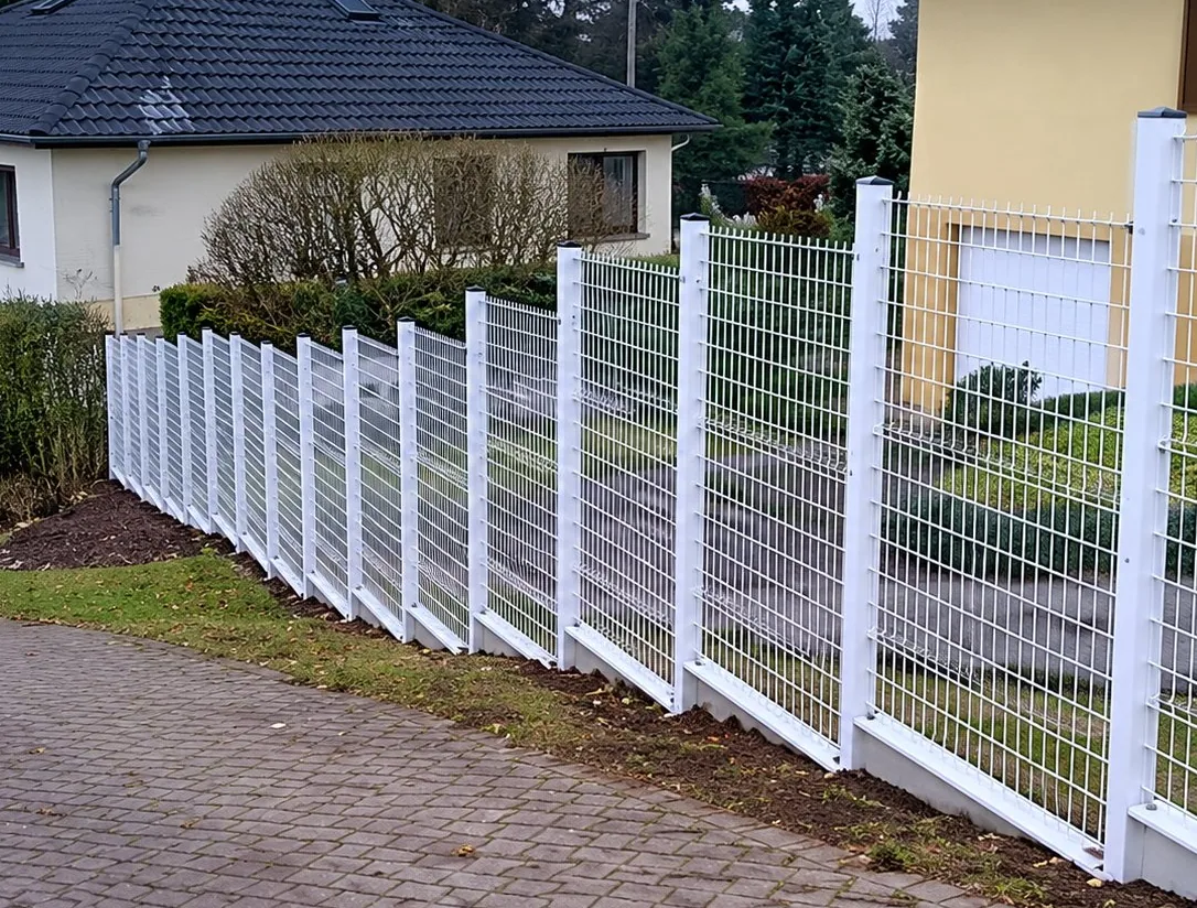 Une clôture en panneaux grillagés blancs longe un trottoir pavé devant une maison aux murs clairs.