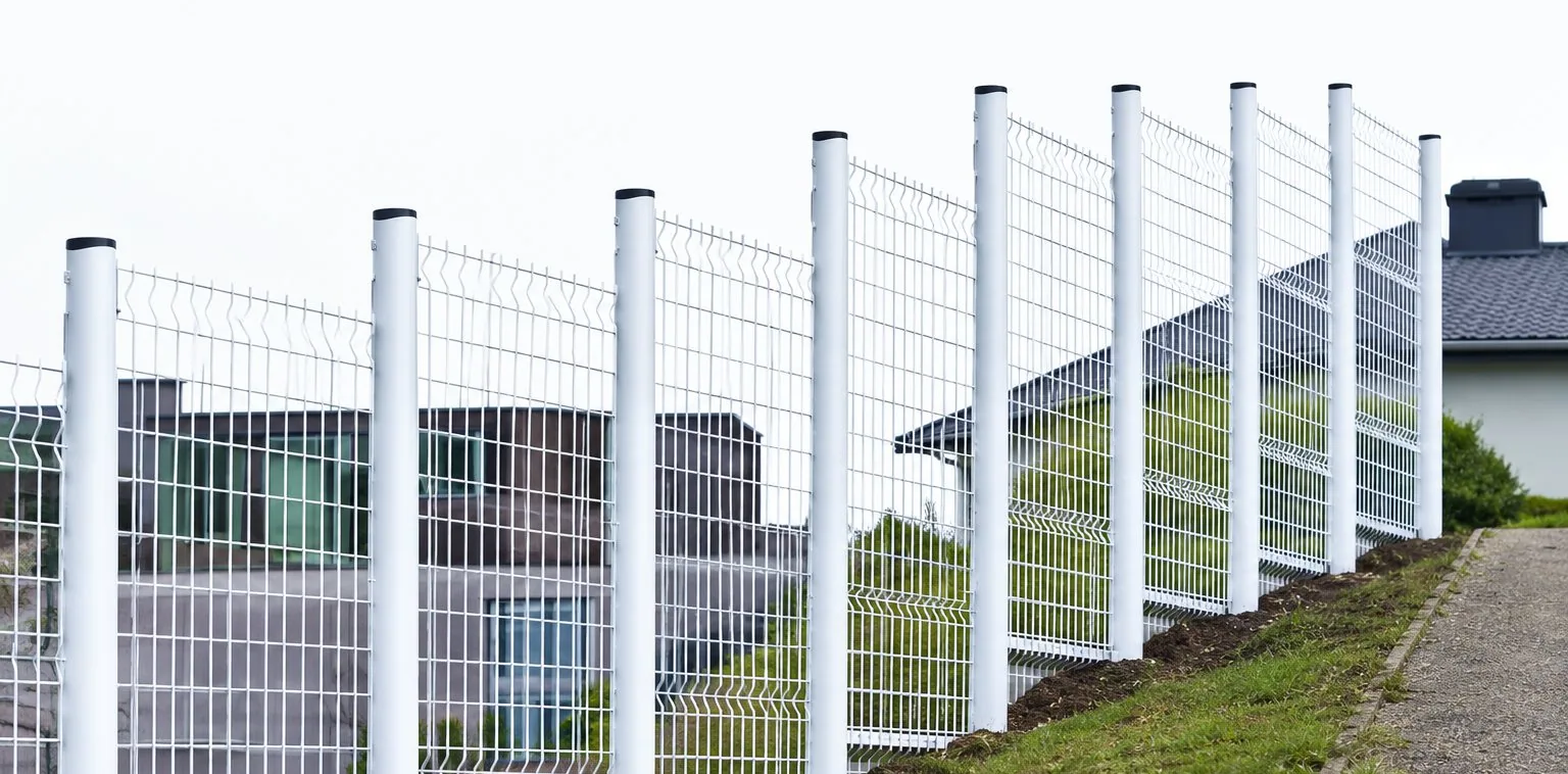 Une clôture blanche à panneaux grillagés longe un talus herbeux, avec une maison en arrière-plan.