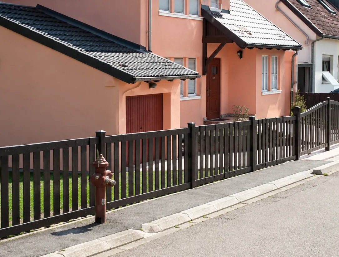 Une clôture noire à barreaux verticaux longe un trottoir devant des maisons aux façades roses et blanches.