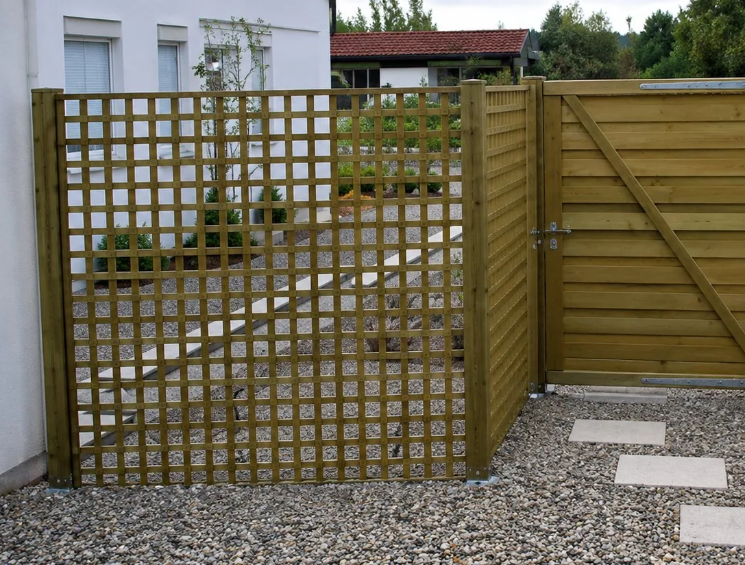 Une clôture et un portail en bois à lattes croisées délimitent une allée en gravier avec des dalles claires, devant une maison.