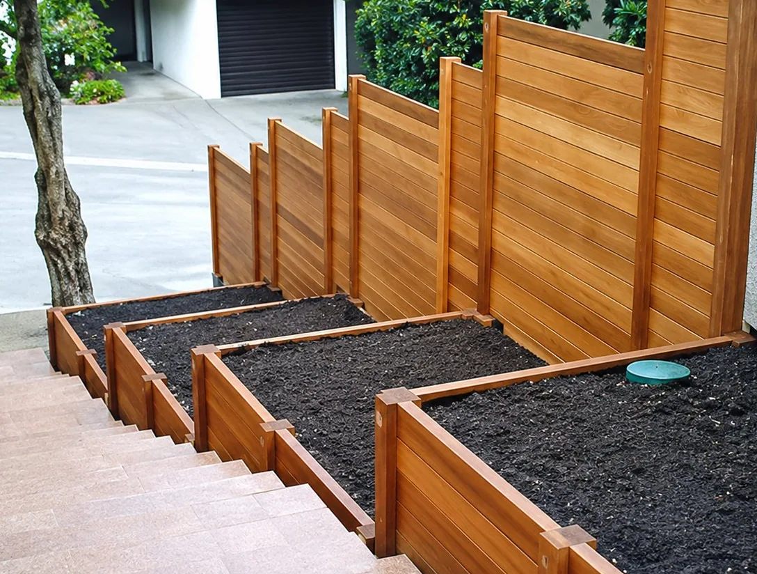 Un escalier extérieur en paliers bordés de jardinières en bois rempli de terre, avec une palissade en bois à droite et un petit arbre à gauche.