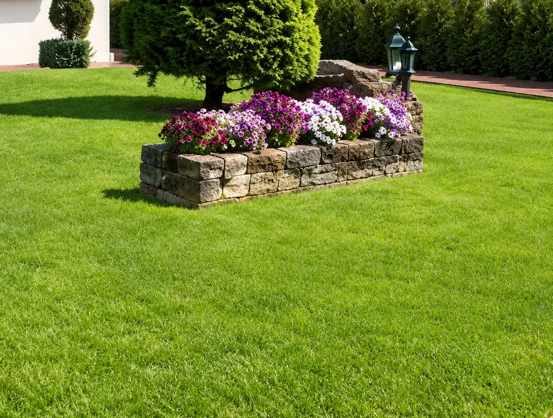 Une jardinière en pierre remplie de fleurs roses et violettes au milieu d'une grande pelouse verte, avec une haie et des arbres en arrière-plan.