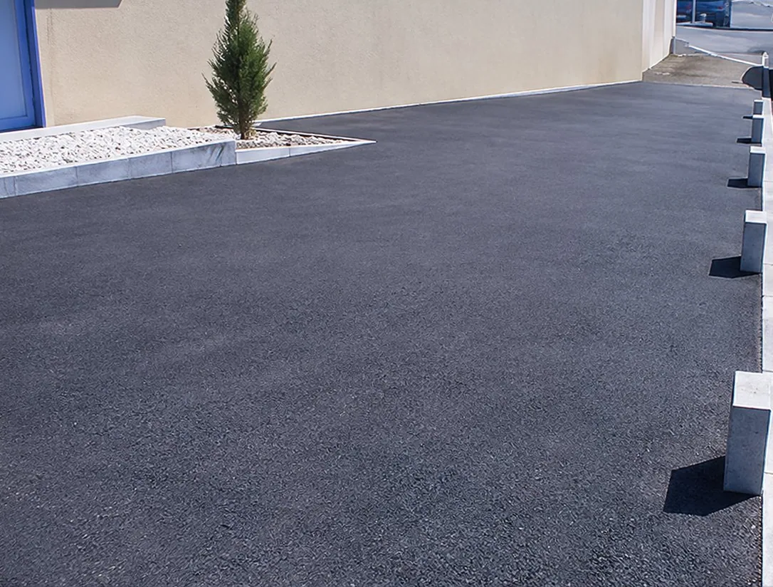 Un parking récemment revêtu d'enrobé noir, bordé de plots et d'une ligne blanche, devant un bâtiment beige avec un petit arbuste.