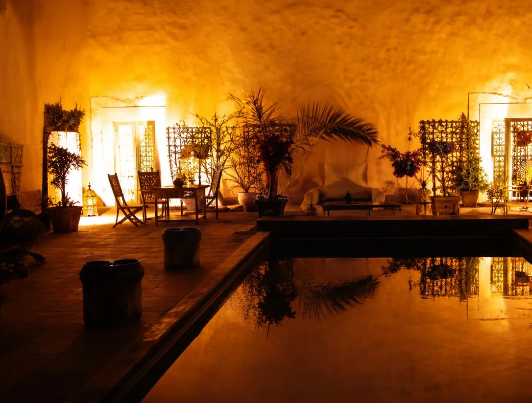 Intérieur d'une salle aux murs de pierre baignée d'une lumière orangée, avec un bassin d'eau au premier plan qui reflète les lumières.