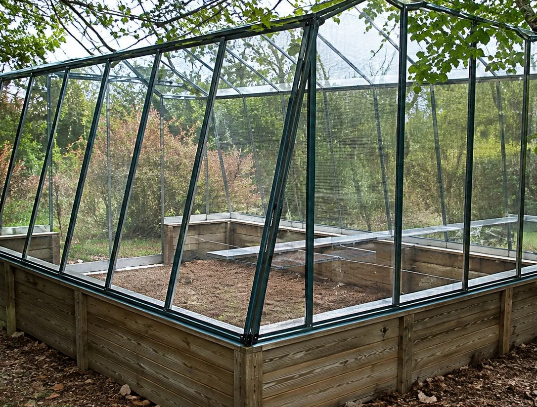 Une serre de jardin aux parois de verre et à l'armature métallique, posée sur un muret en bois, entourée d'arbres.