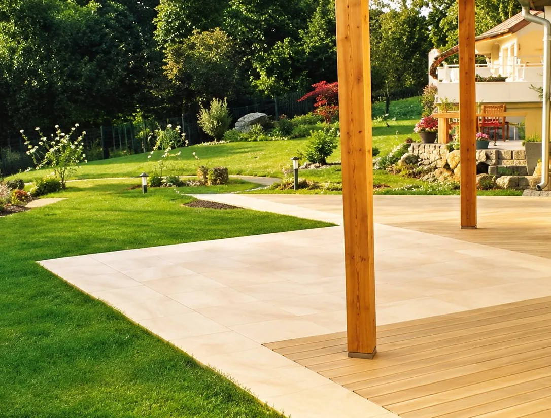 Une terrasse aménagée avec des dalles claires et un platelage en bois, soutenue par des poteaux en bois, au milieu d'un jardin avec pelouse verte et arbres.