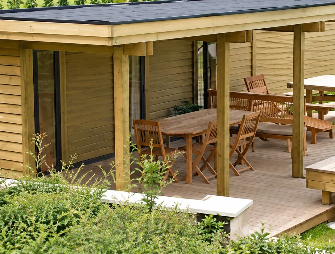Une terrasse en bois sous un auvent avec une table et des chaises, entourée de pelouse et de verdure.