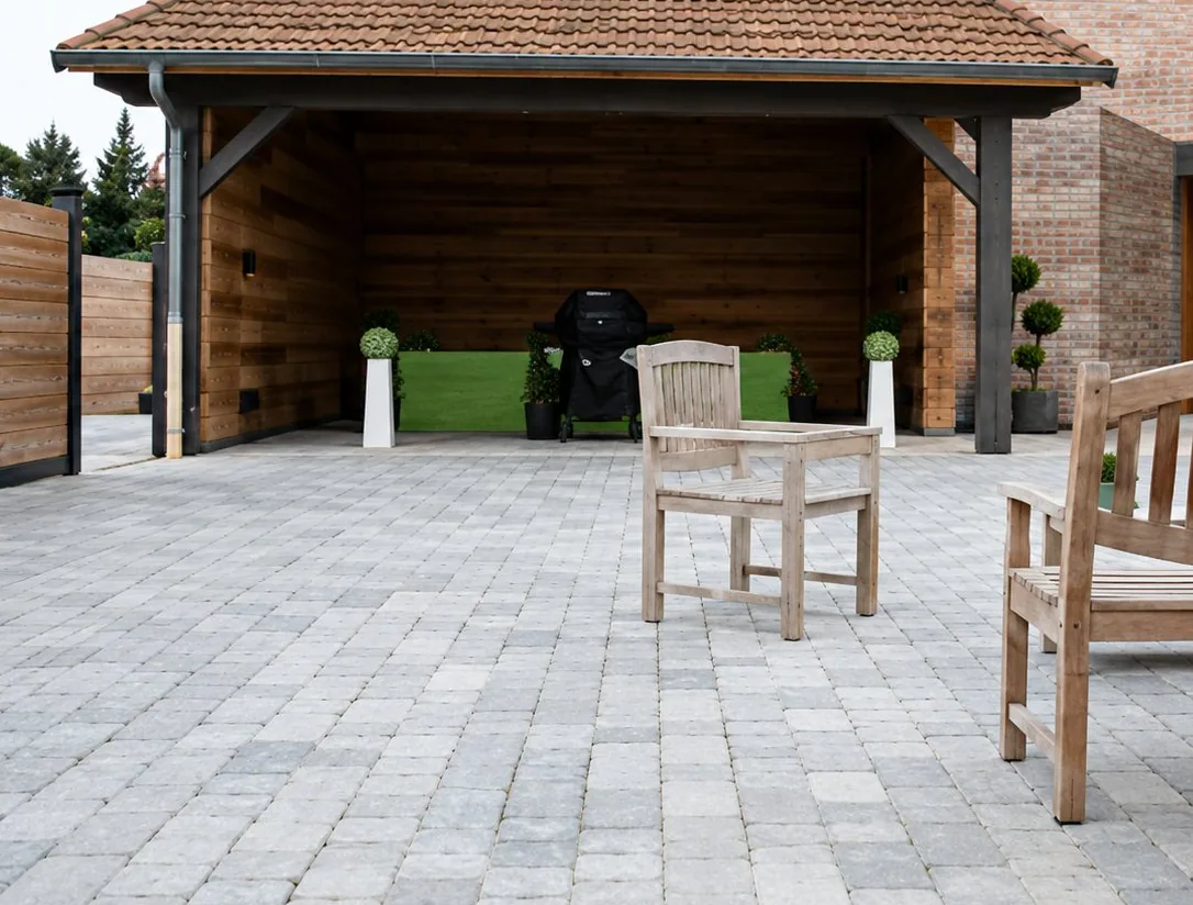 Une terrasse récemment pavée de dalles grises devant un abri couvert en bois, avec une chaise blanche et un banc en bois au premier plan.