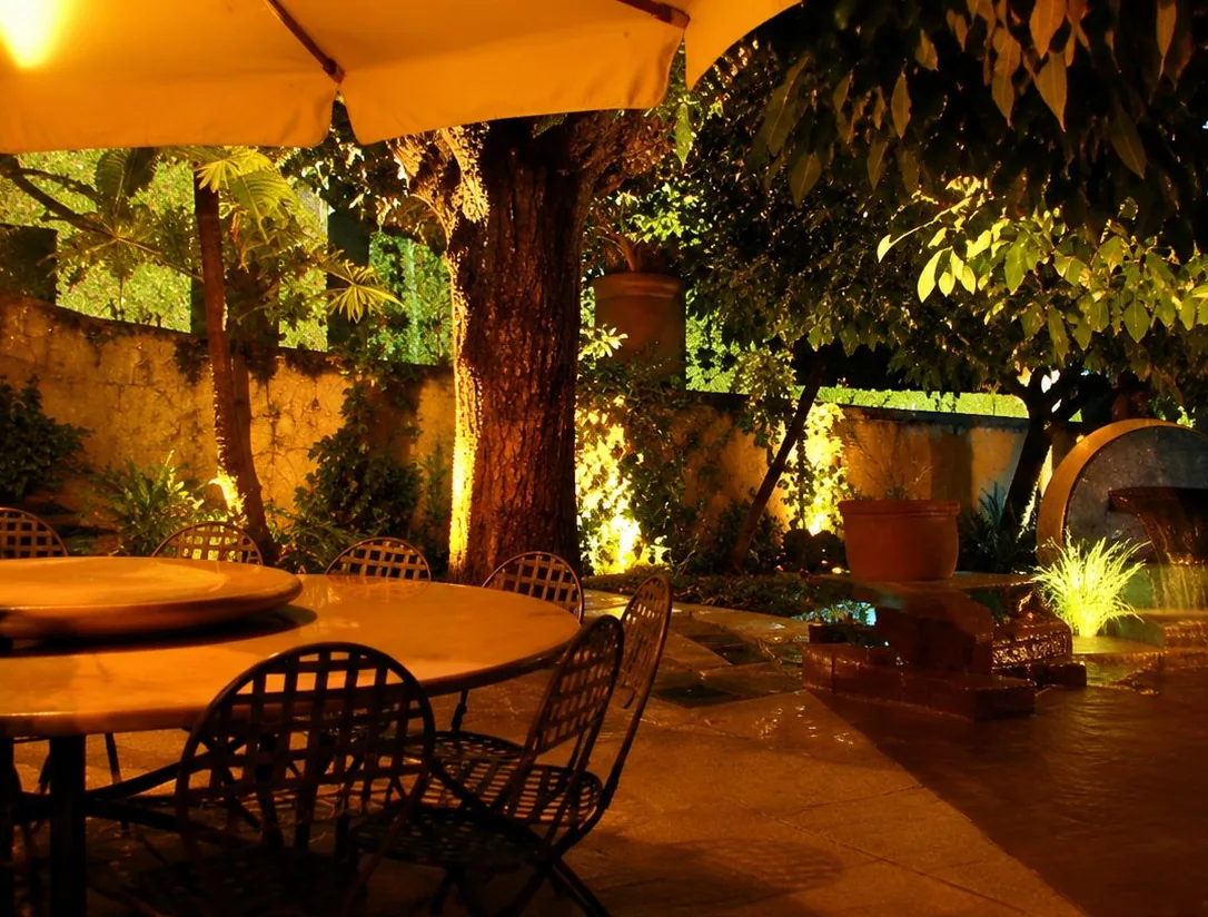 Une terrasse extérieure éclairée en soirée, avec des tables rondes, des chaises en fer forgé, un parasol et de la végétation illuminée.